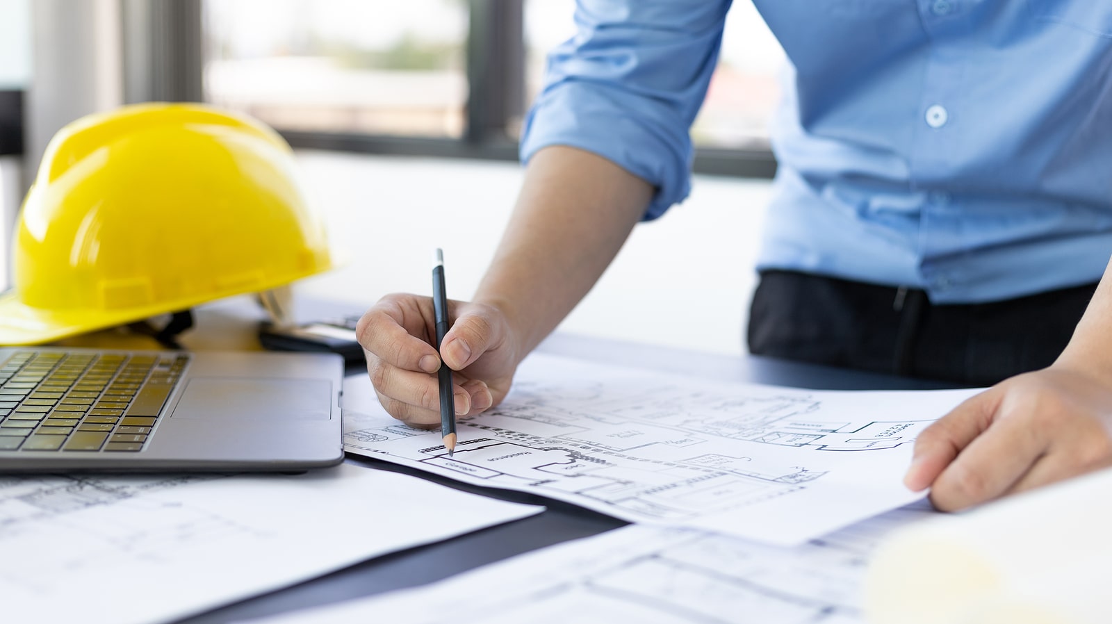 A person creating an architectural blueprint with a hard hat in the background.